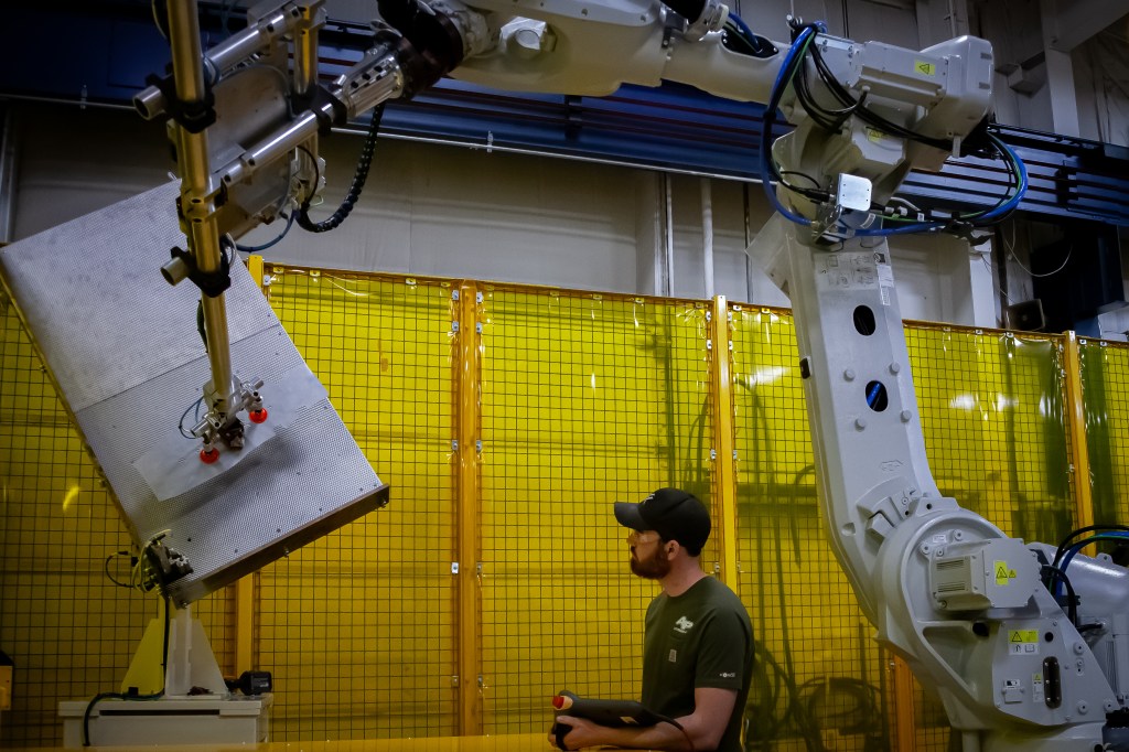 Industrial robot lifting a large flat part with custom end-of-arm tooling.